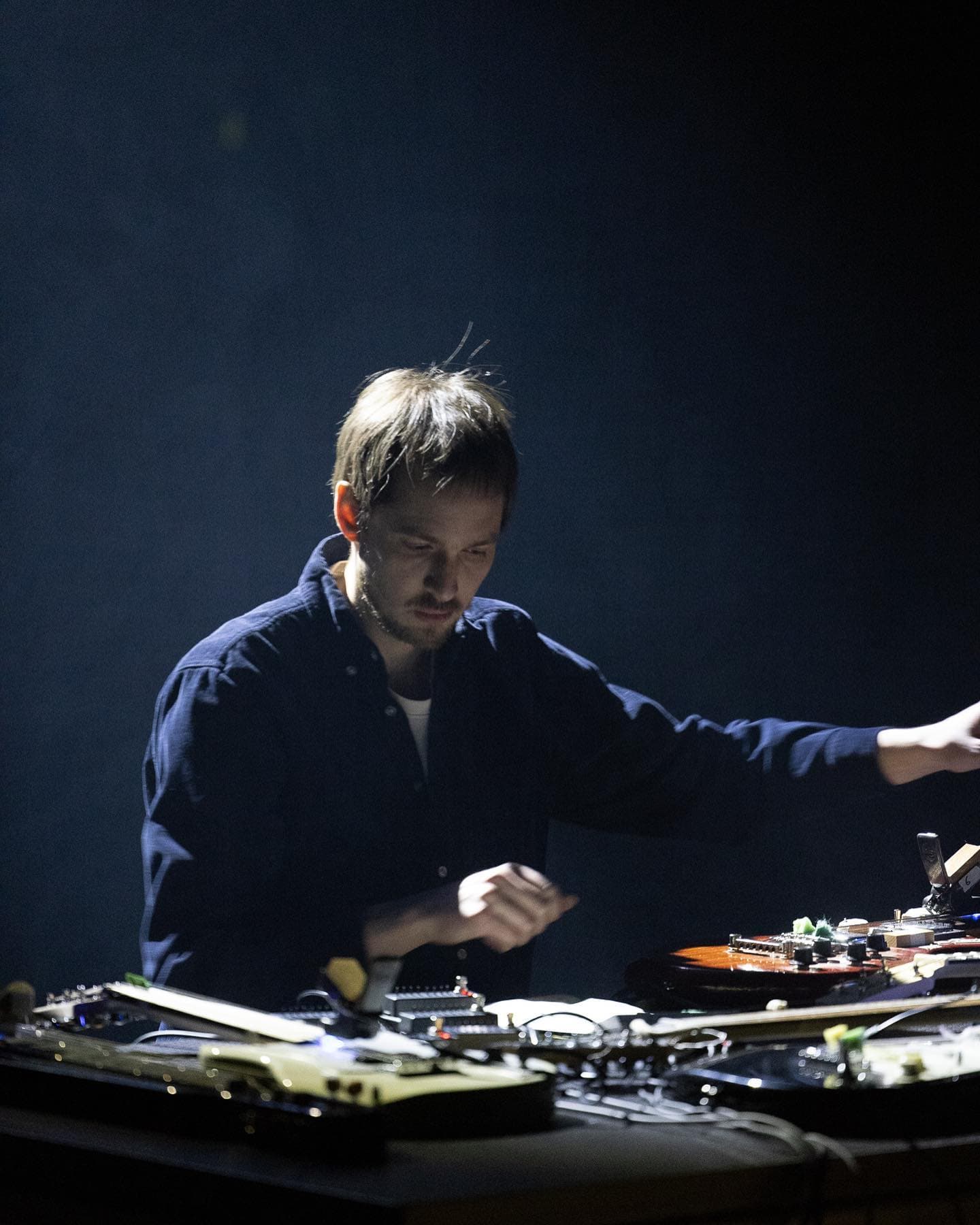 A person is performing with electronic equipment and instruments on a dimly lit stage.
