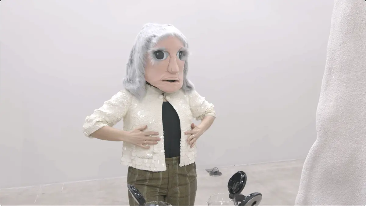 A person wearing a large mask with exaggerated facial features and a white jacket stands in a room.