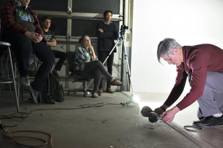 A person is crouching on a floor adjusting a device while several others sit or stand watching in a dimly lit room.