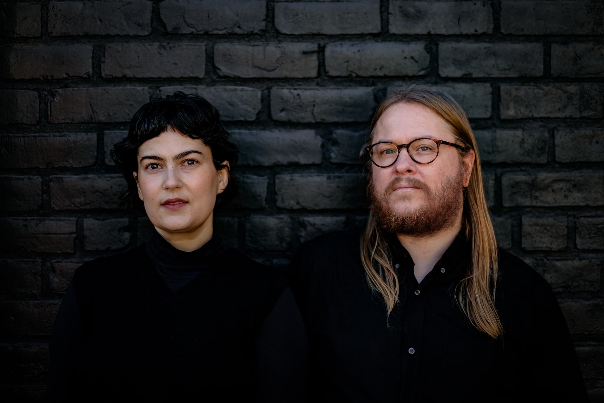 Two people with dark clothing stand against a dark brick wall, one with short hair and the other with long hair and glasses.