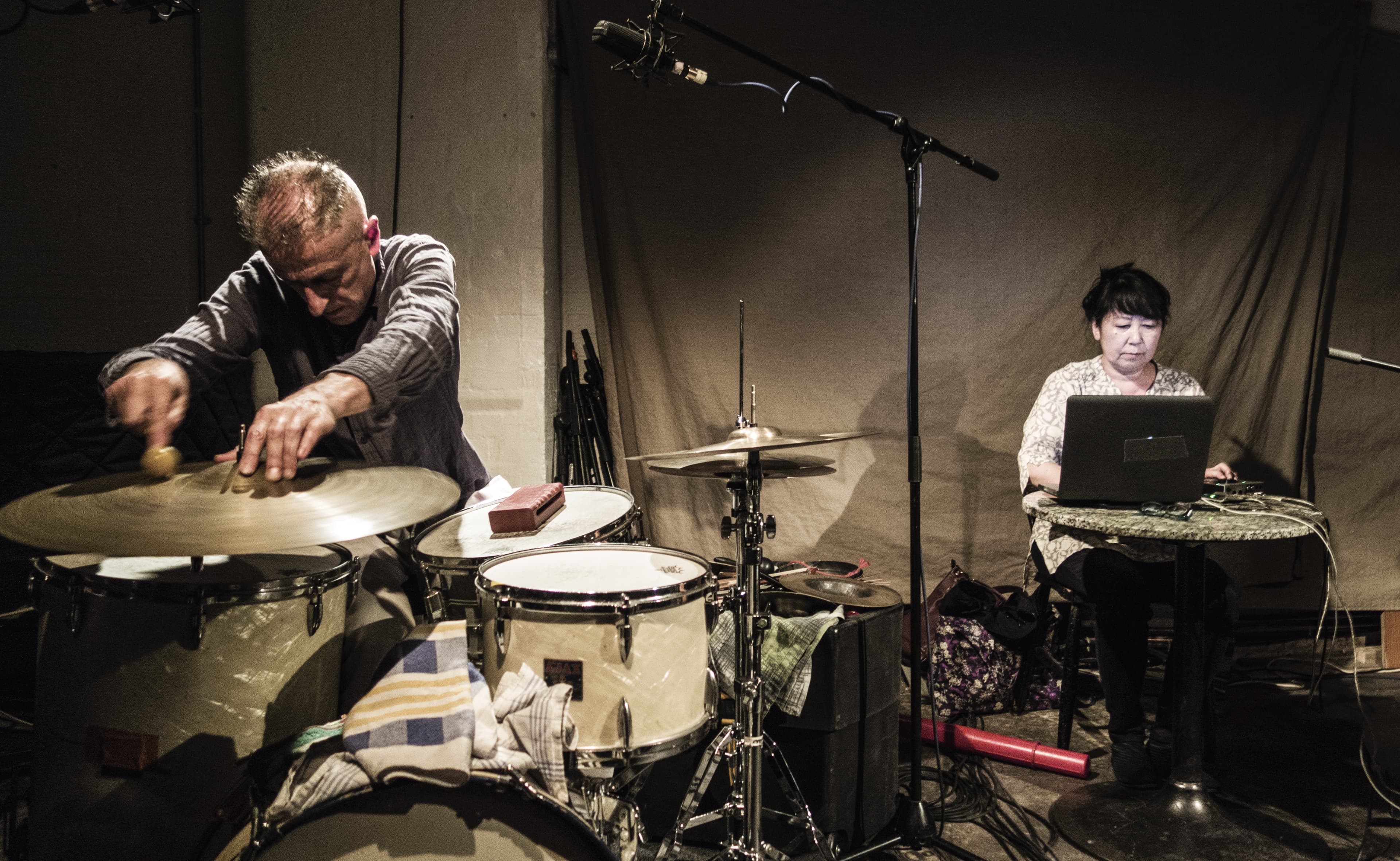 A person is playing drums while another performs using a laptop at a table, with microphones set up nearby.