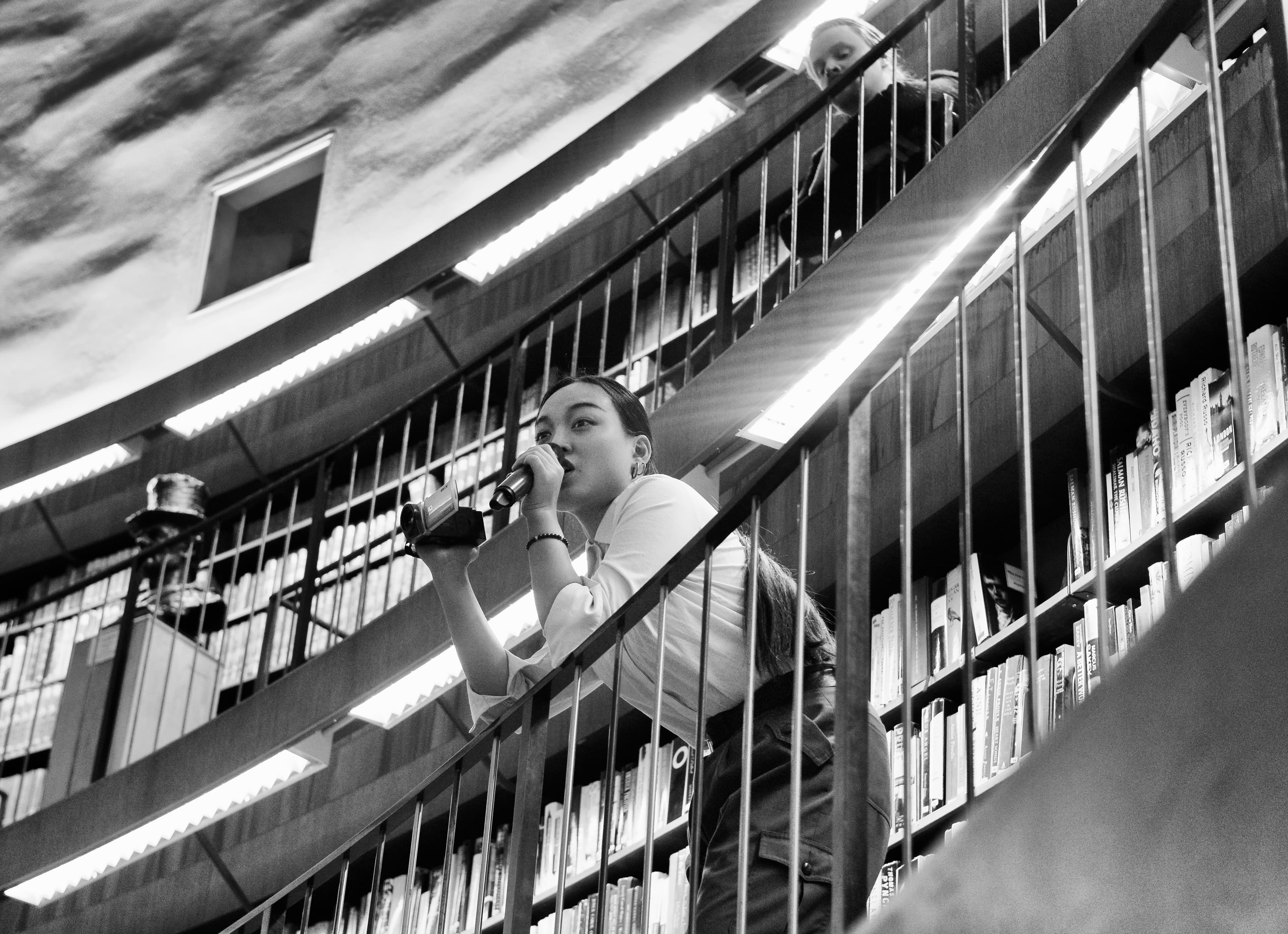 A woman is speaking into a microphone on a staircase in a library, with bookshelves in the background.