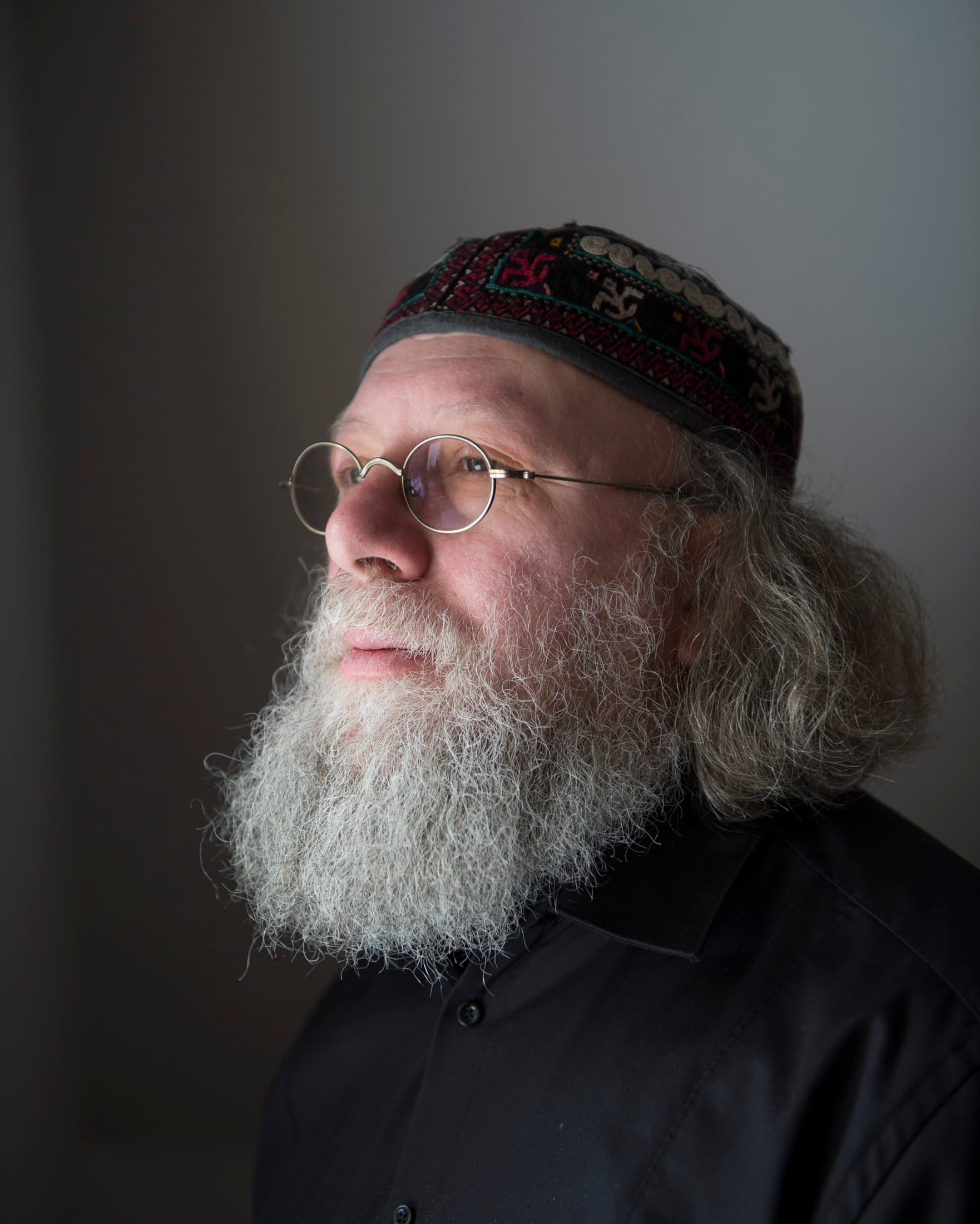 A man with glasses and a long white beard is wearing a patterned cap and a black shirt, looking to the side.