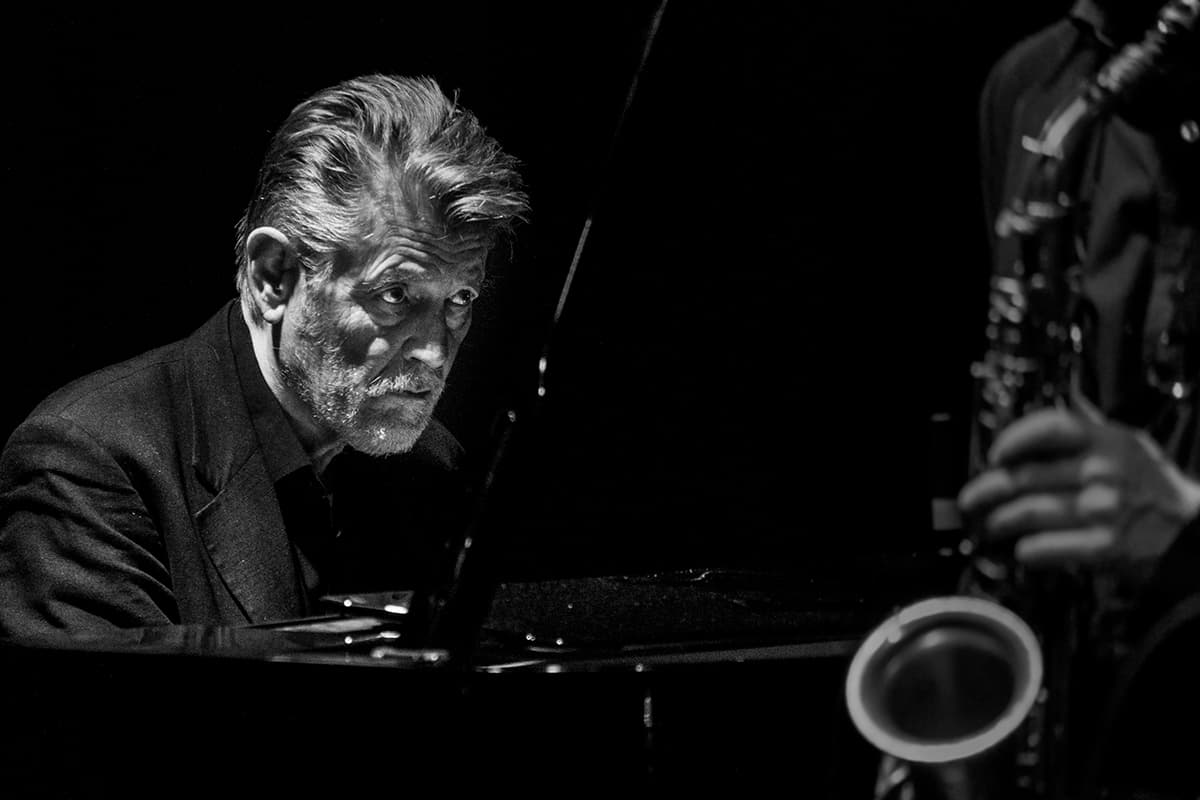 A man playing piano in a dimly lit setting, with part of a saxophone and its player visible.