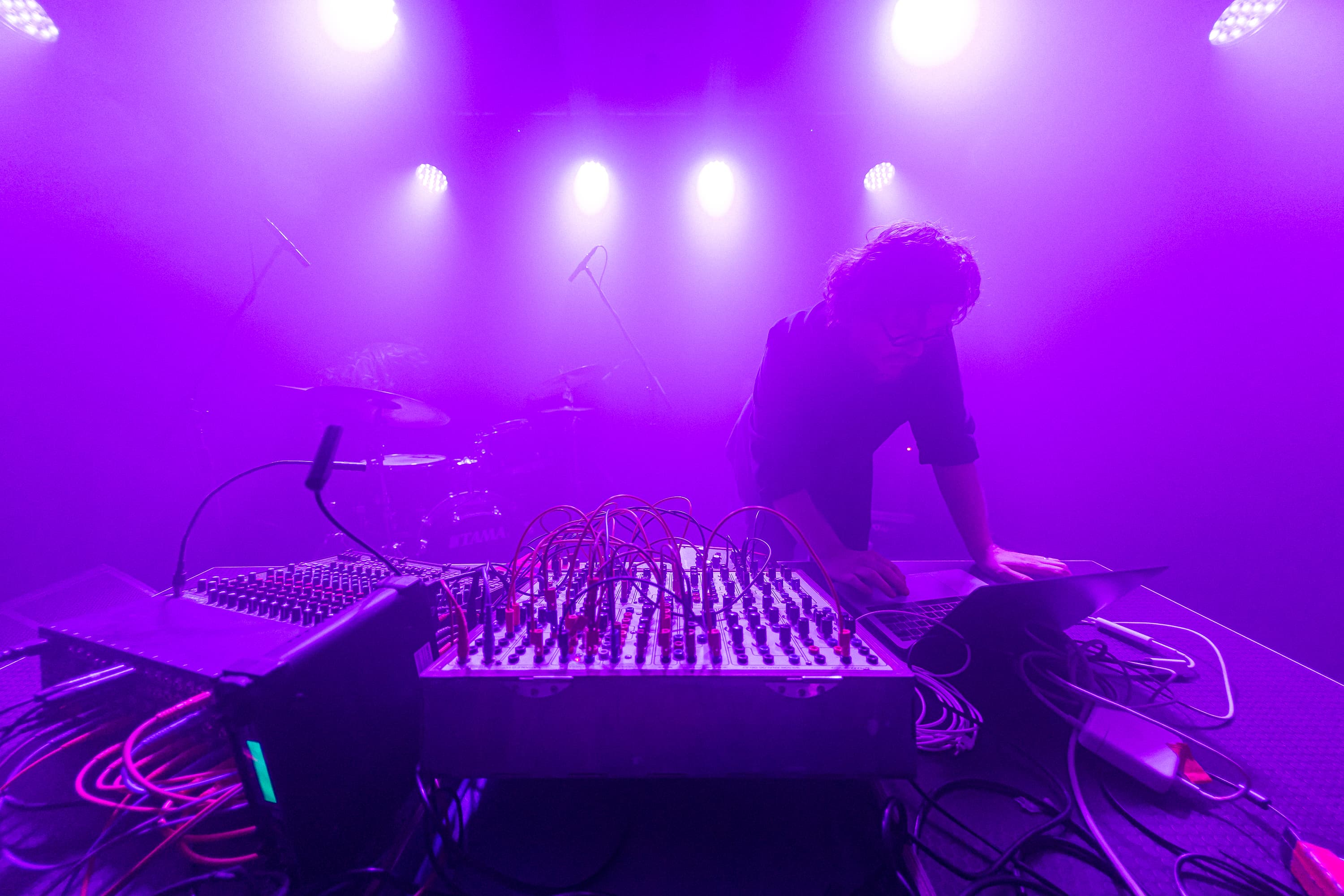 A person is operating electronic music equipment on stage, surrounded by purple lighting and musical instruments.