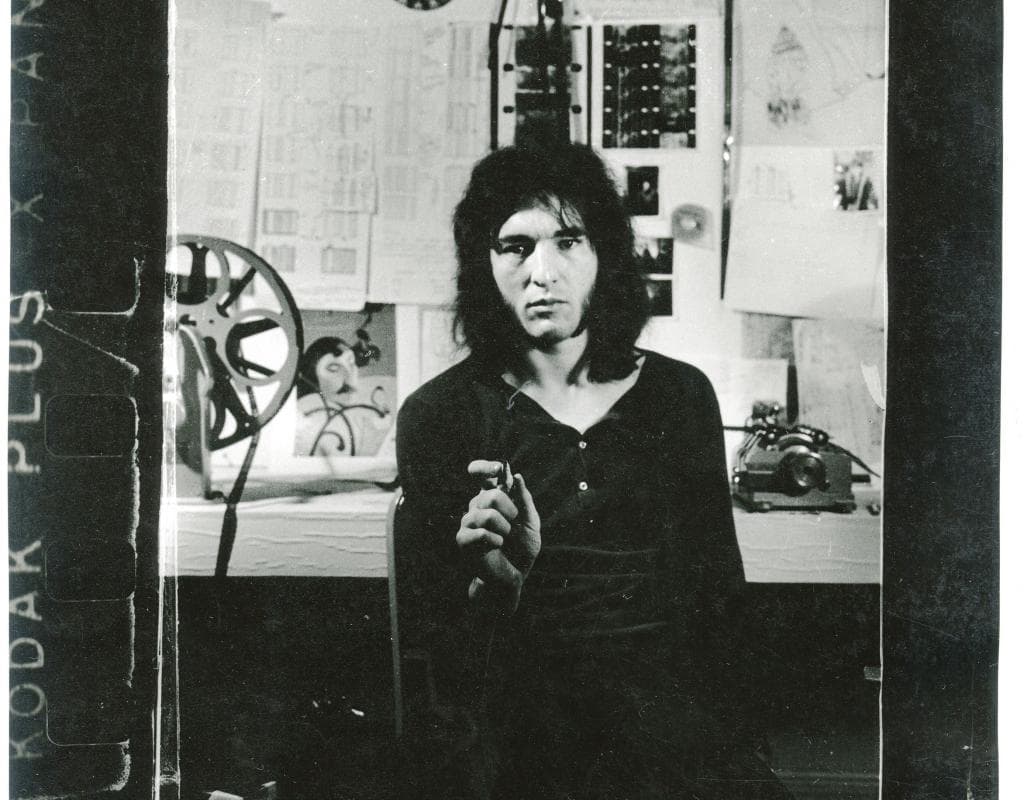 A person with long hair is seated in a room with film reels, drawings, and equipment in the background.