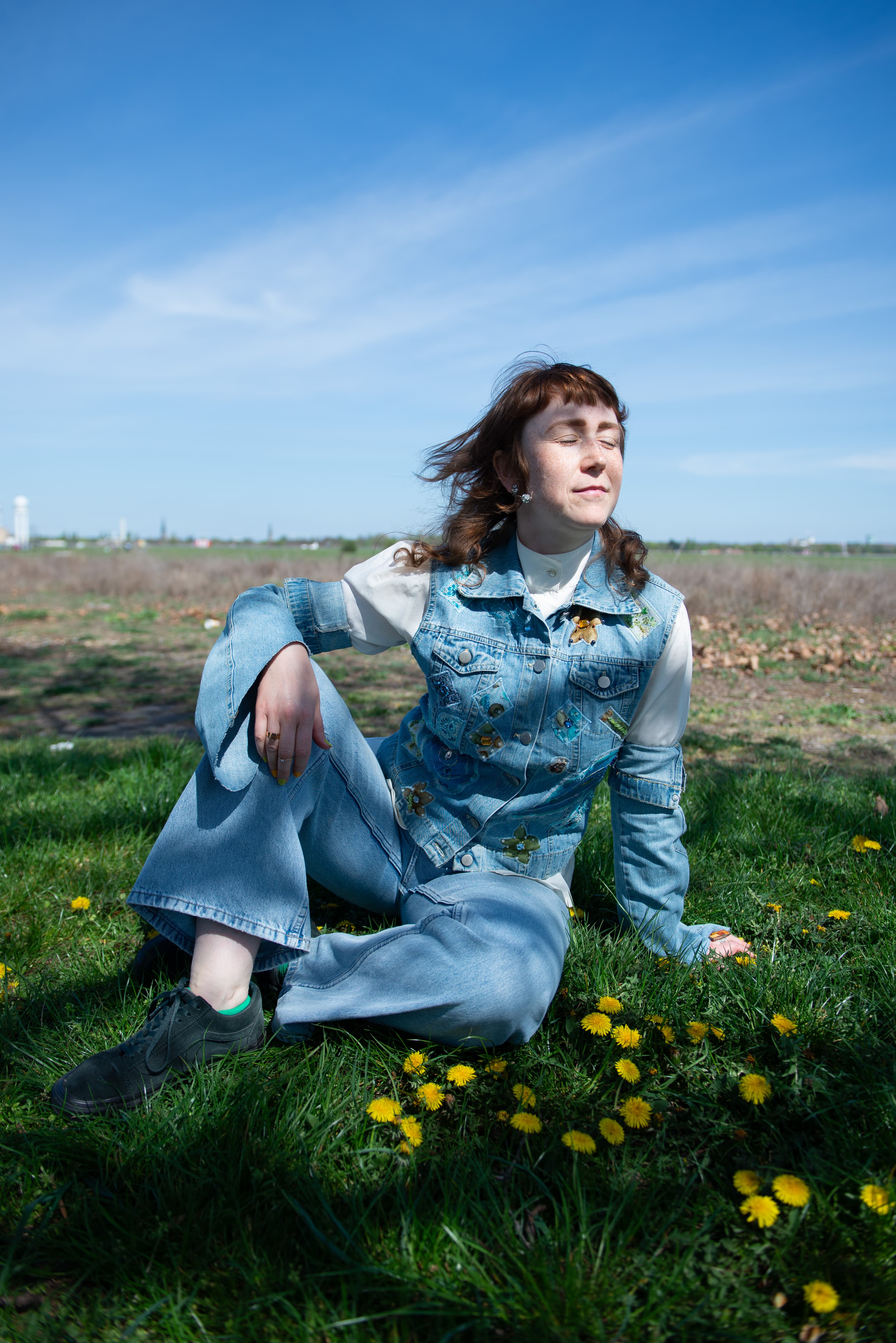A person wearing a denim outfit sits on grass surrounded by yellow flowers, eyes closed, under a clear blue sky.