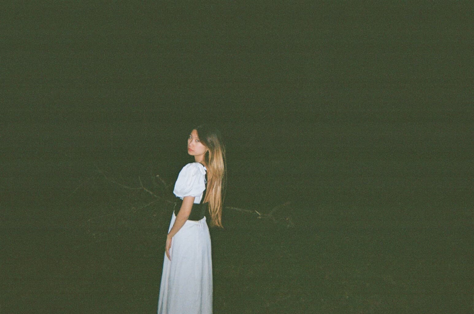 A woman in a white dress stands in a dark outdoor setting, looking over her shoulder.