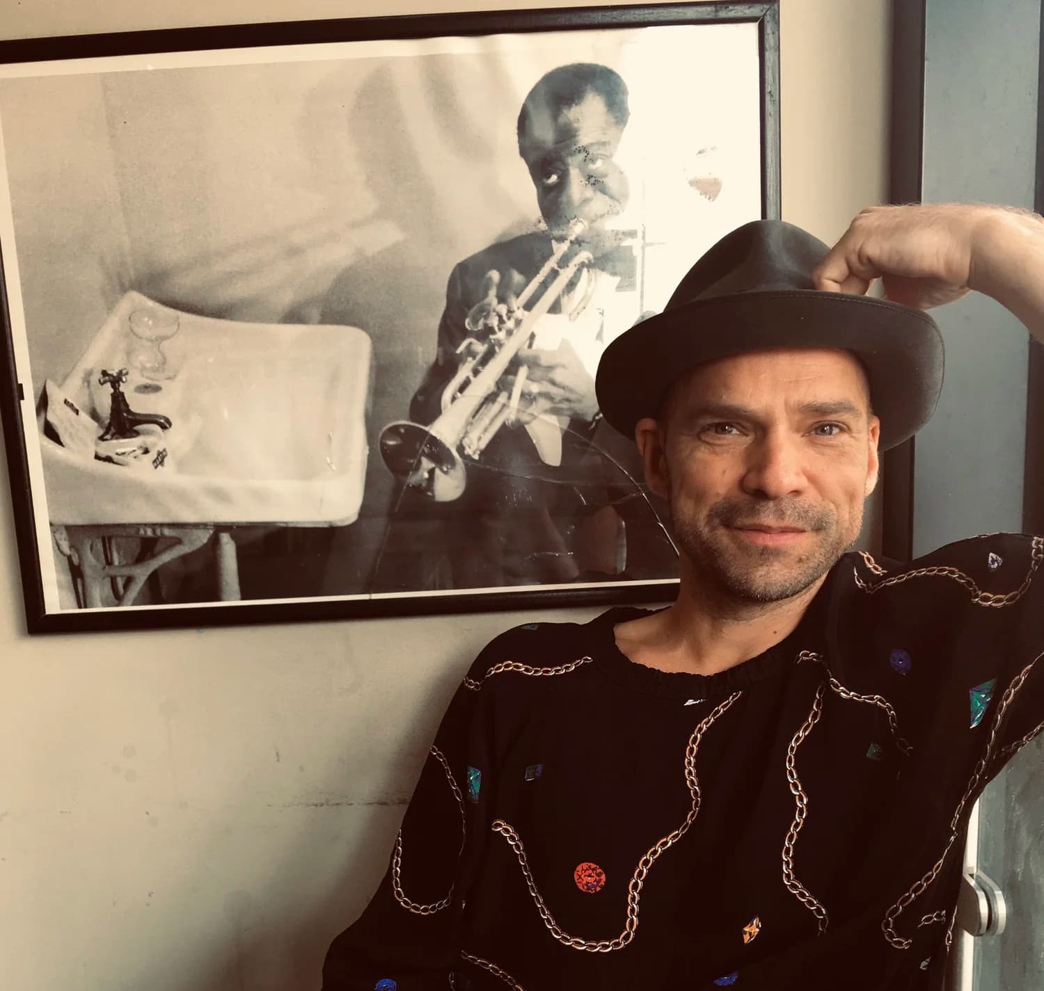 A person wearing a hat poses in front of a framed black-and-white photo of a louis armstrong playing the trumpet.