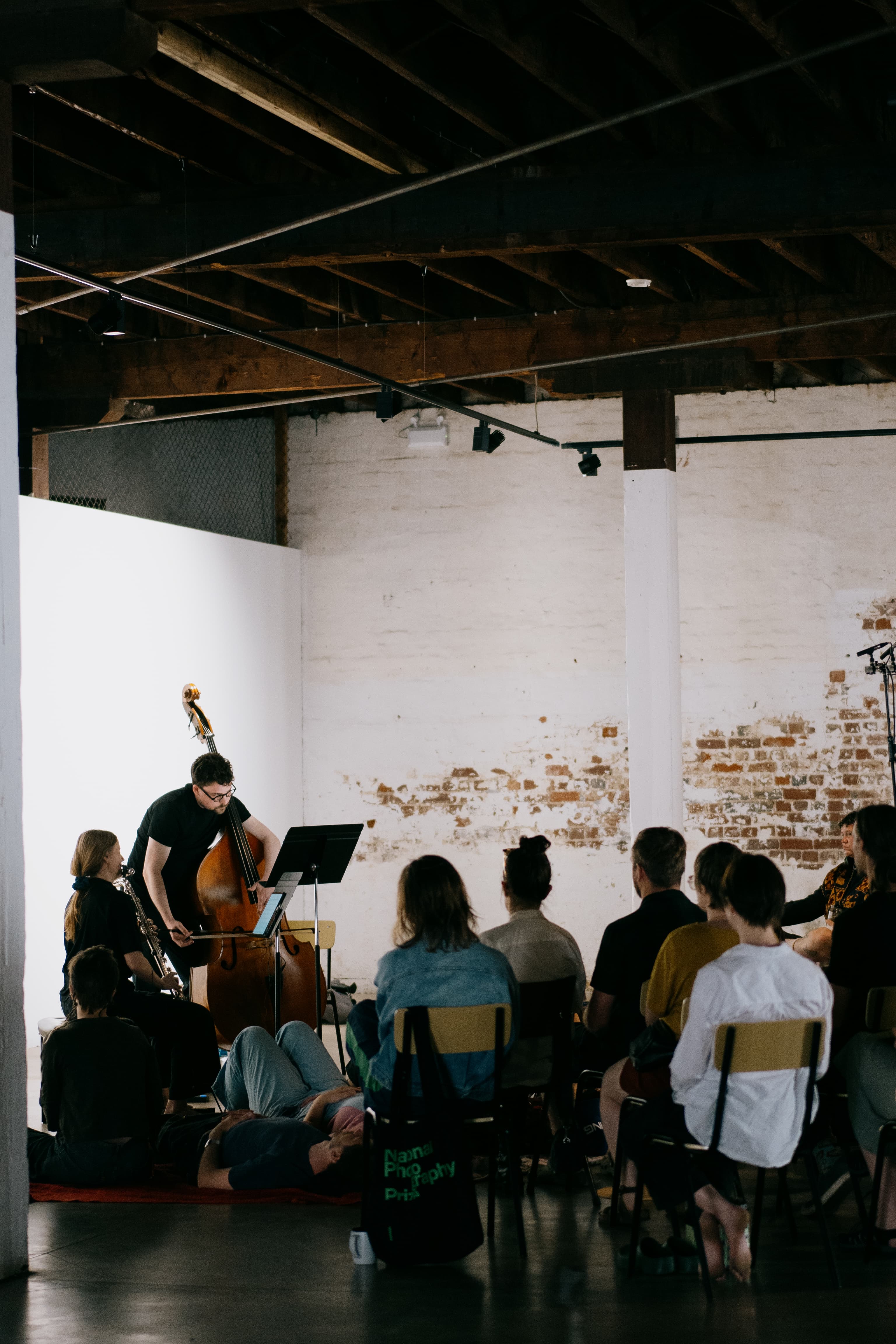 A small audience watches two musicians playing a string instrument and a quarter-tone flute in a rustic indoor setting.