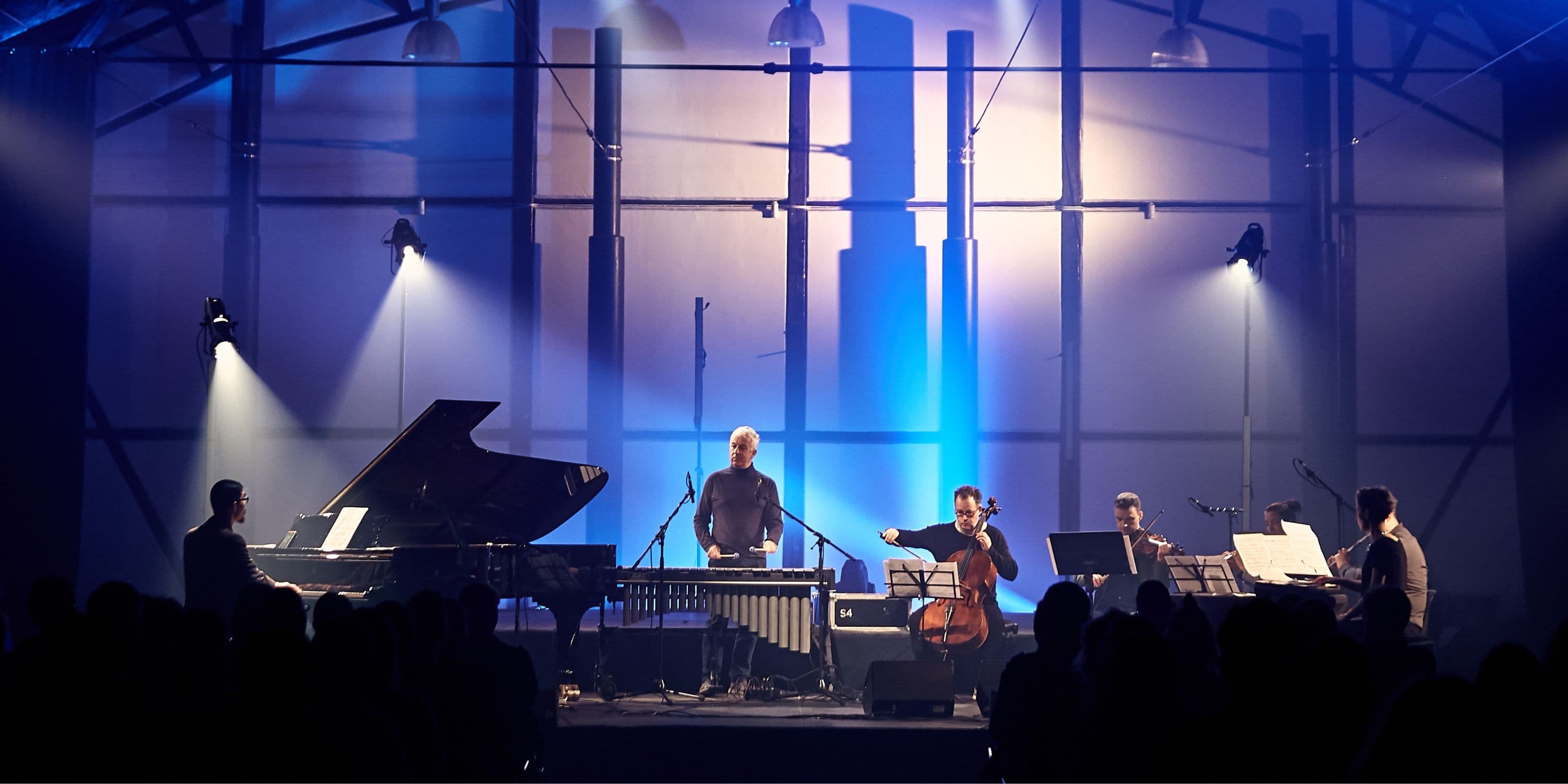 A group of musicians is performing on stage, illuminated by spotlights, with a piano, string instruments, and percussion.