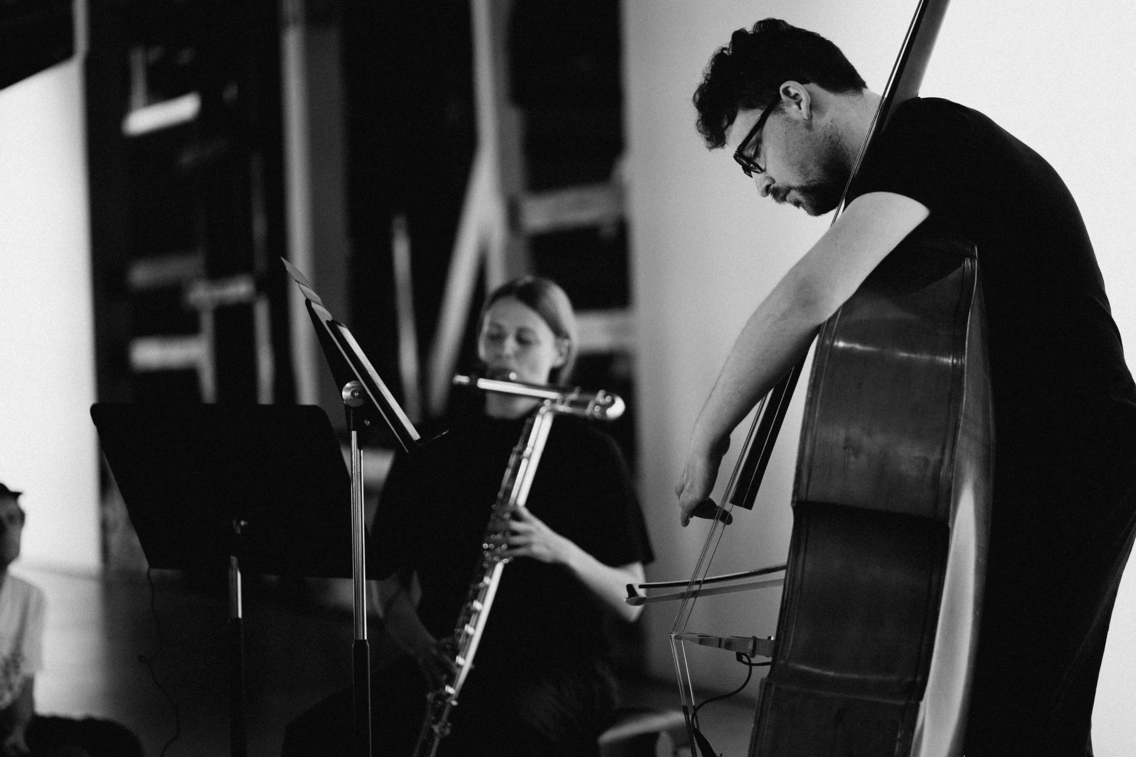 A black and white scene of two musicians performing, one playing a bass and the other a bass clarinet, with music stands in front of them.