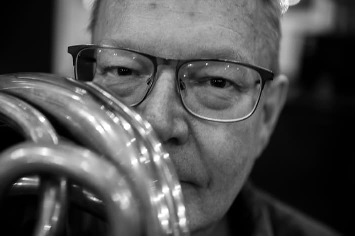 A person wearing glasses is playing a brass instrument (tuba), captured in close-up and black and white.