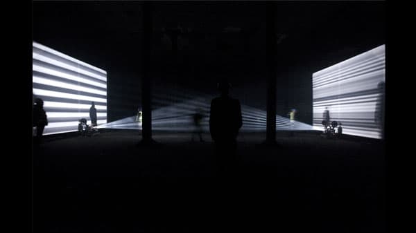 A dark room features two large screens with horizontal white stripes, with silhouettes of people standing and moving in the space.