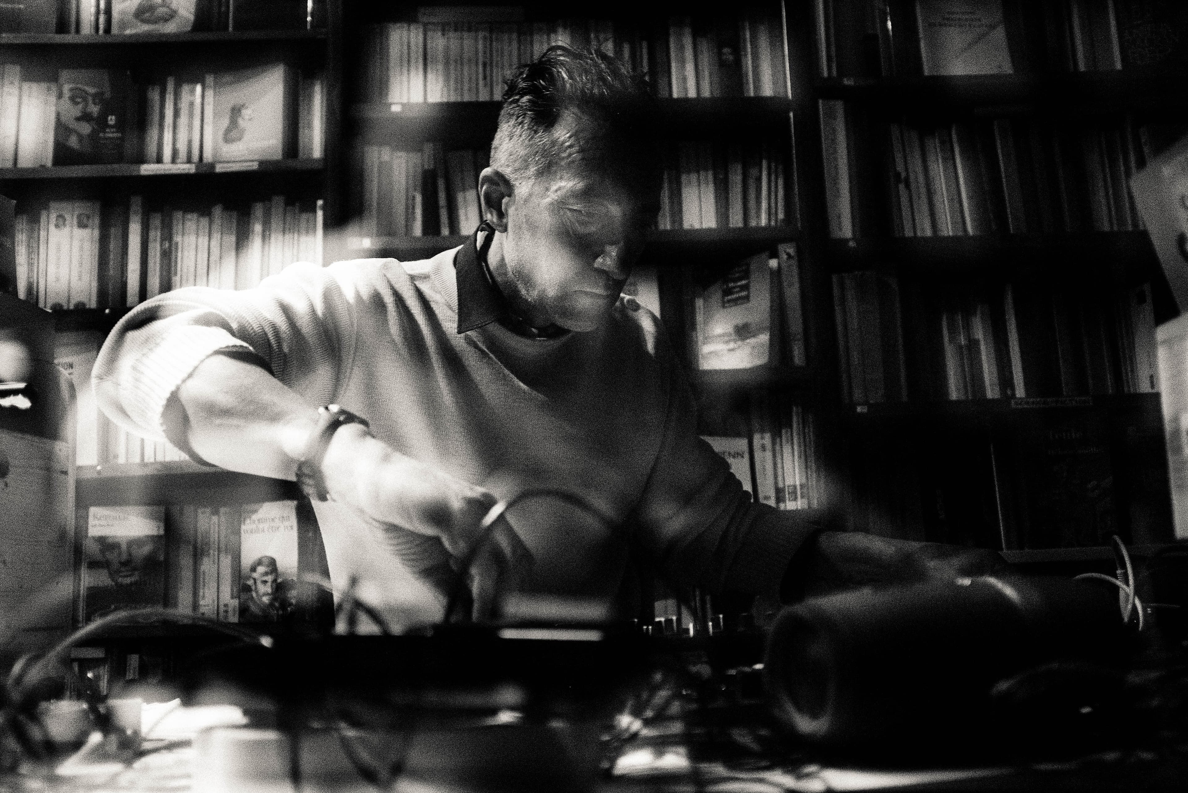 A person is working at a cluttered desk in a dimly lit room filled with bookshelves.