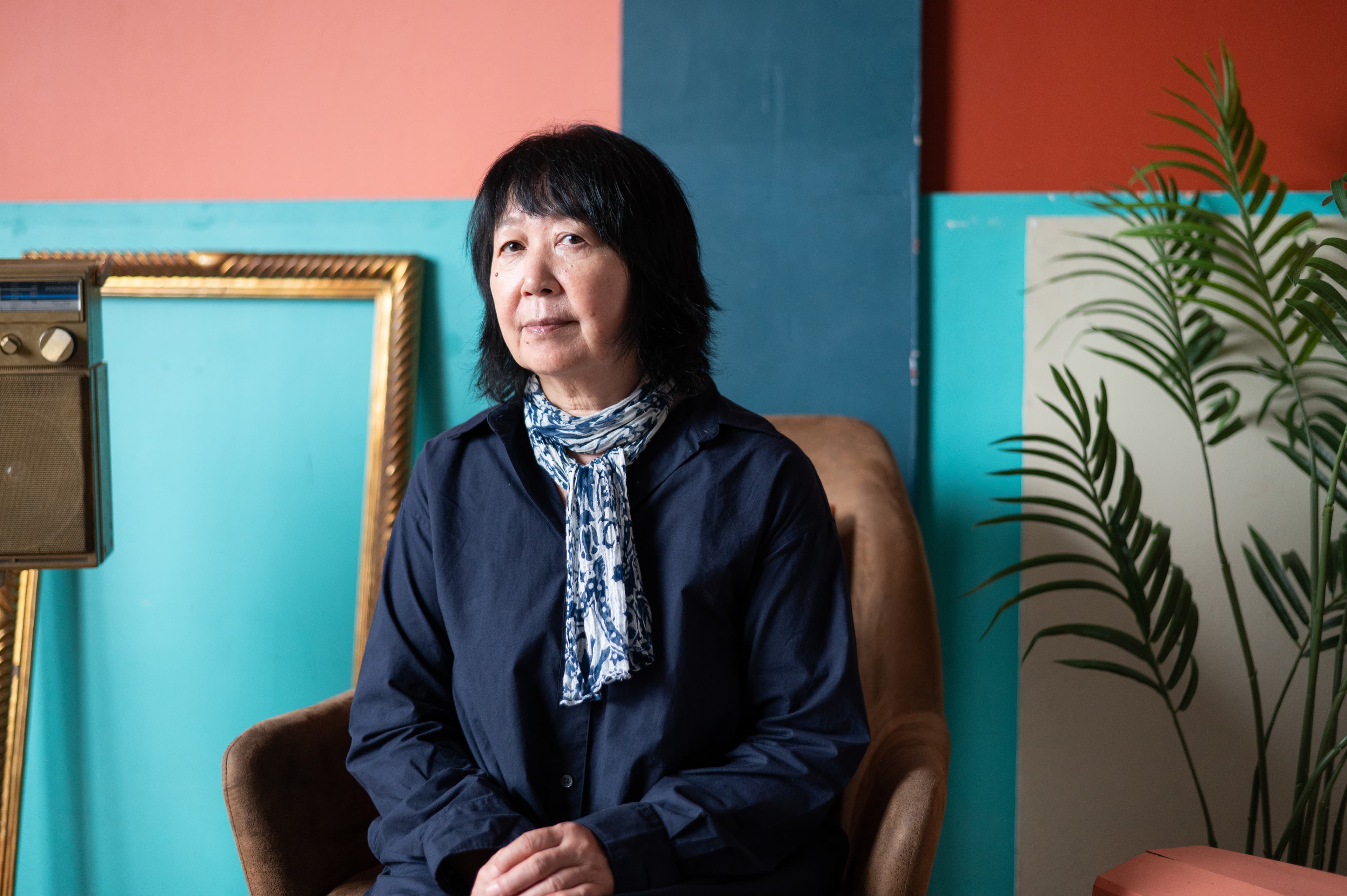 A woman with black hair, wearing a blue scarf and dark shirt, sits in a chair against a colorful wall, with a vintage radio and plant nearby.