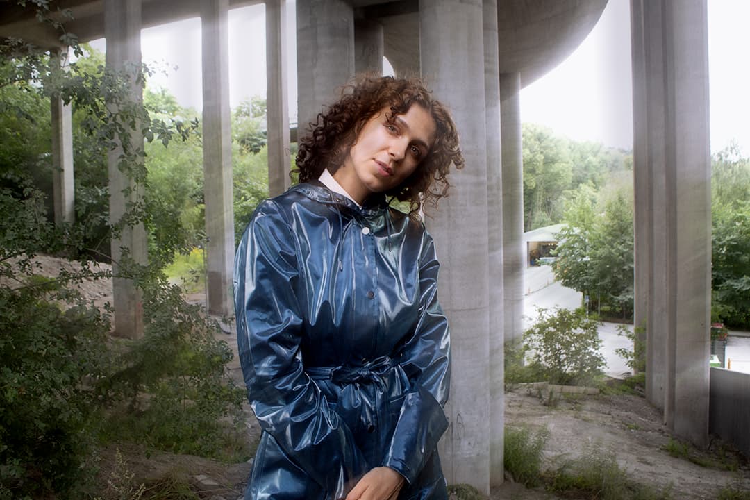 A person in a blue raincoat stands under a bridge, surrounded by greenery.