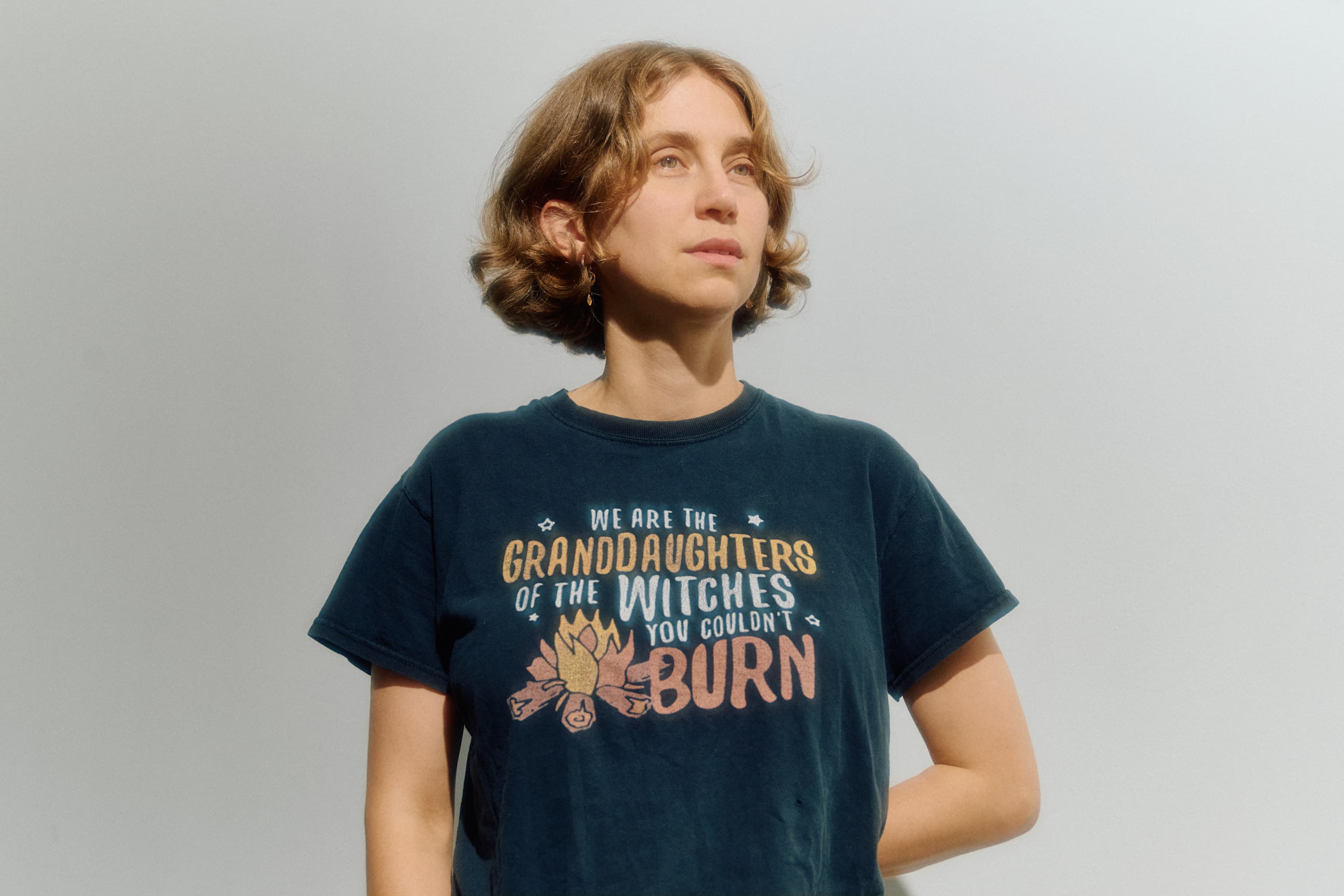 A person with short hair is wearing a dark T-shirt that reads, "We are the granddaughters of the witches you couldn't burn."