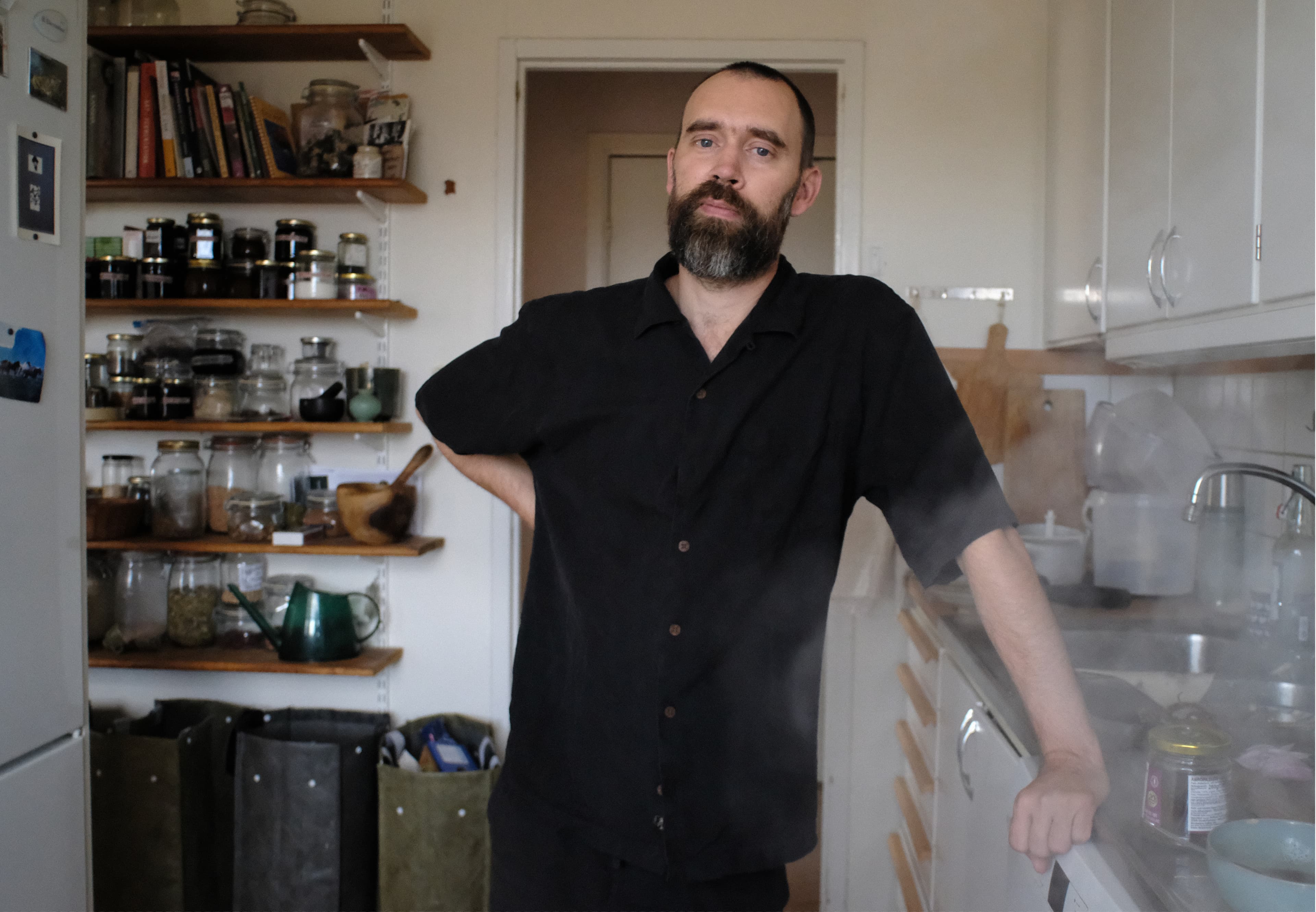 A man with a beard stands in a kitchen with shelves of jars and books behind him.