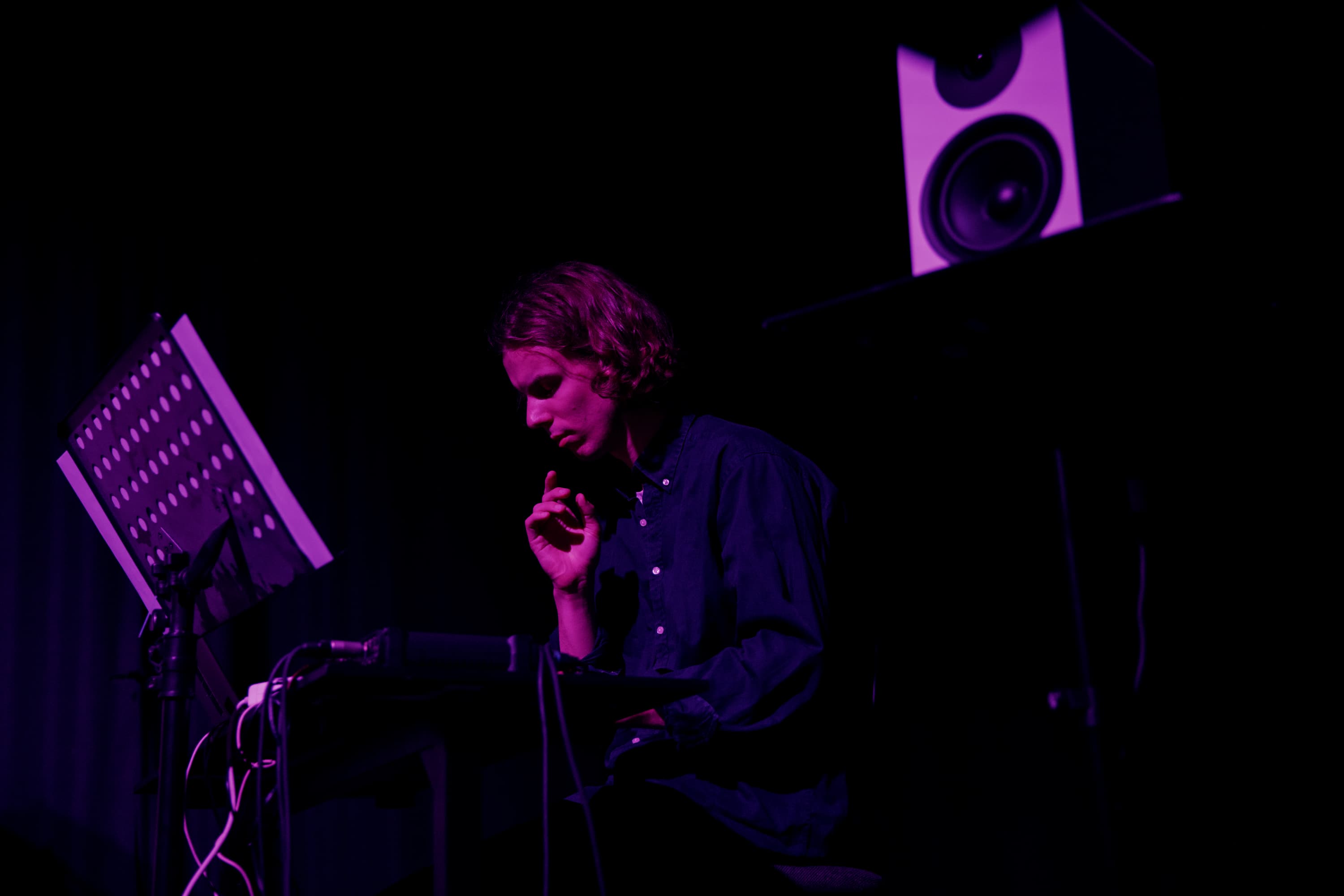 A person is sitting in a dimly lit room, focusing on a musical setup with a music stand and speaker nearby.
