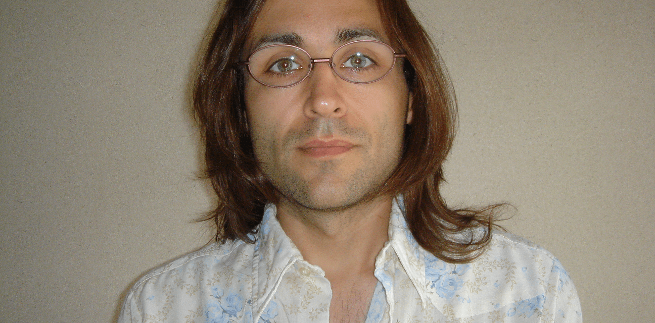 A person with long brown hair and glasses is wearing a floral patterned shirt and looking directly at the camera.