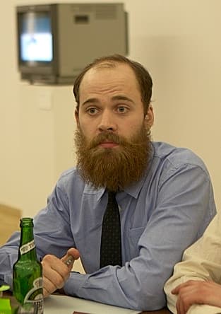 A bearded man in a blue shirt and tie sits at a table with a bottle in front of him.