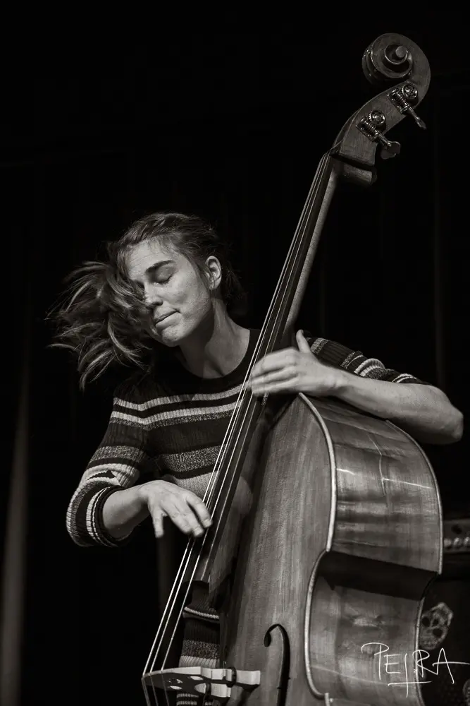 A person passionately plays a double bass, eyes closed, with hair moving as if in motion.