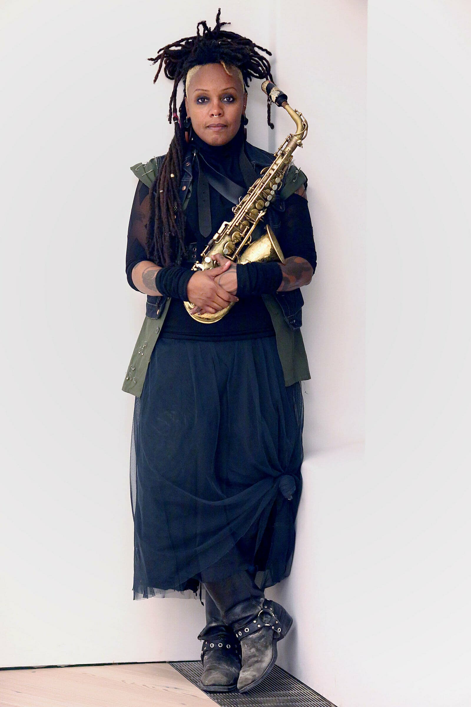 A person with dreadlocks is leaning against a wall, holding a saxophone and wearing a dark outfit with boots.