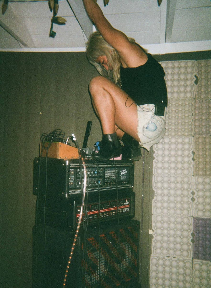 A person with long hair is crouching on top of audio equipment, holding onto the ceiling in a room with acoustic foam on the walls.