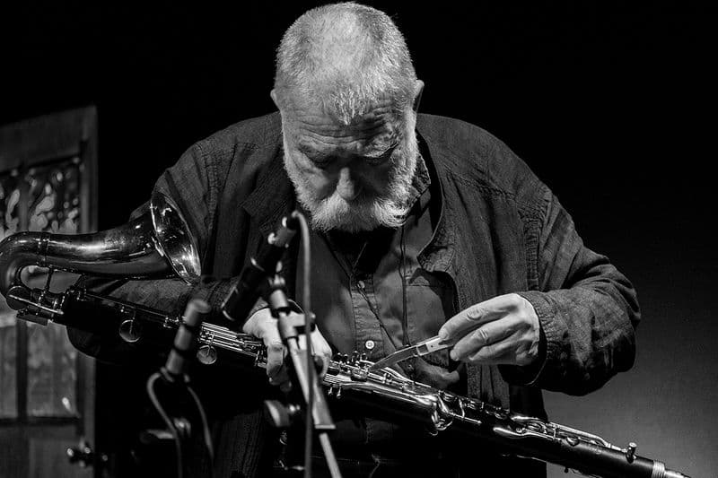 An elderly man with a beard is adjusting his bass clarinet with a small foldable knife.