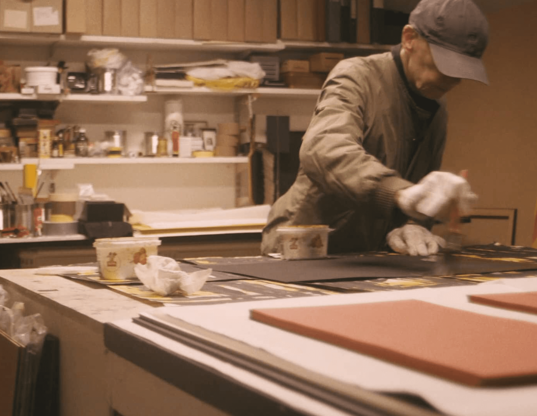 A person wearing a cap and gloves is working on a project at a cluttered workstation with various supplies and tools.
