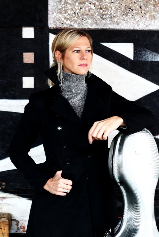 A person with blonde hair in a dark coat stands beside a silver instrument case against a geometric, textured background.