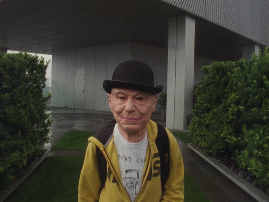 A young person with a serious expression stands in a park with buildings in the background.