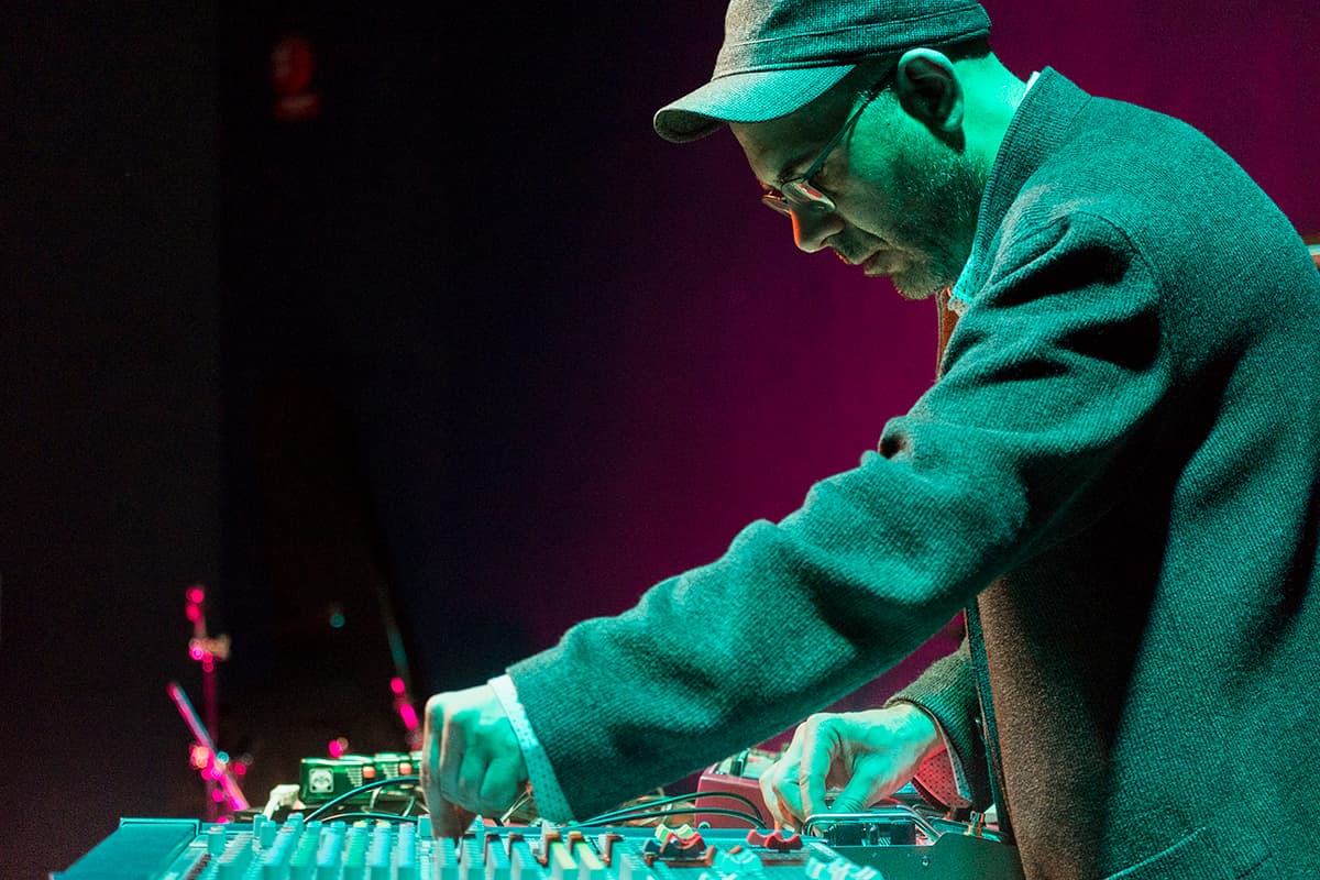 A person wearing a cap and glasses operates a sound mixer under stage lighting.