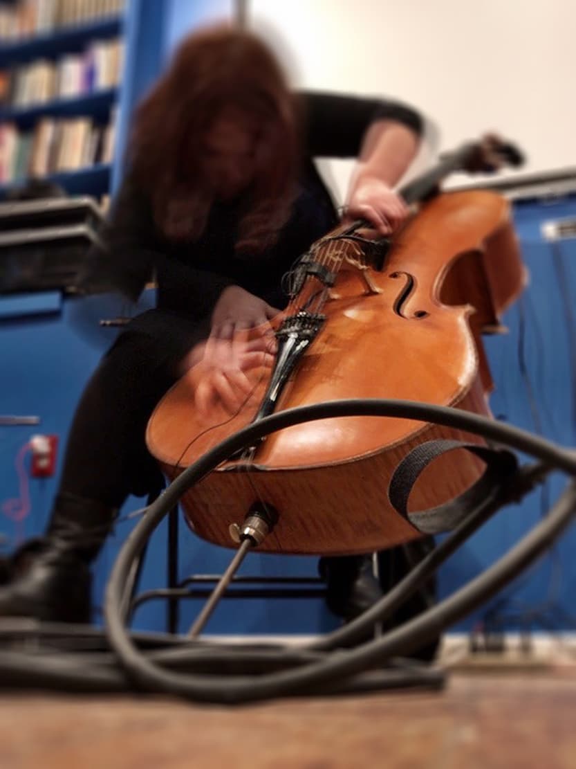 A person is playing a cello with cables on the floor, set against a background of bookshelves.