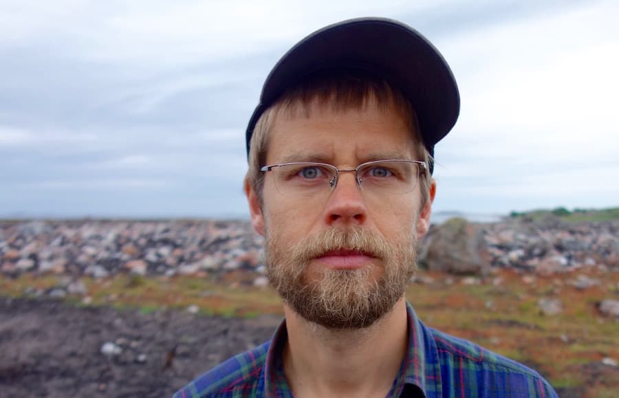 A bearded man wearing glasses and a cap stands outdoors in a rocky landscape.