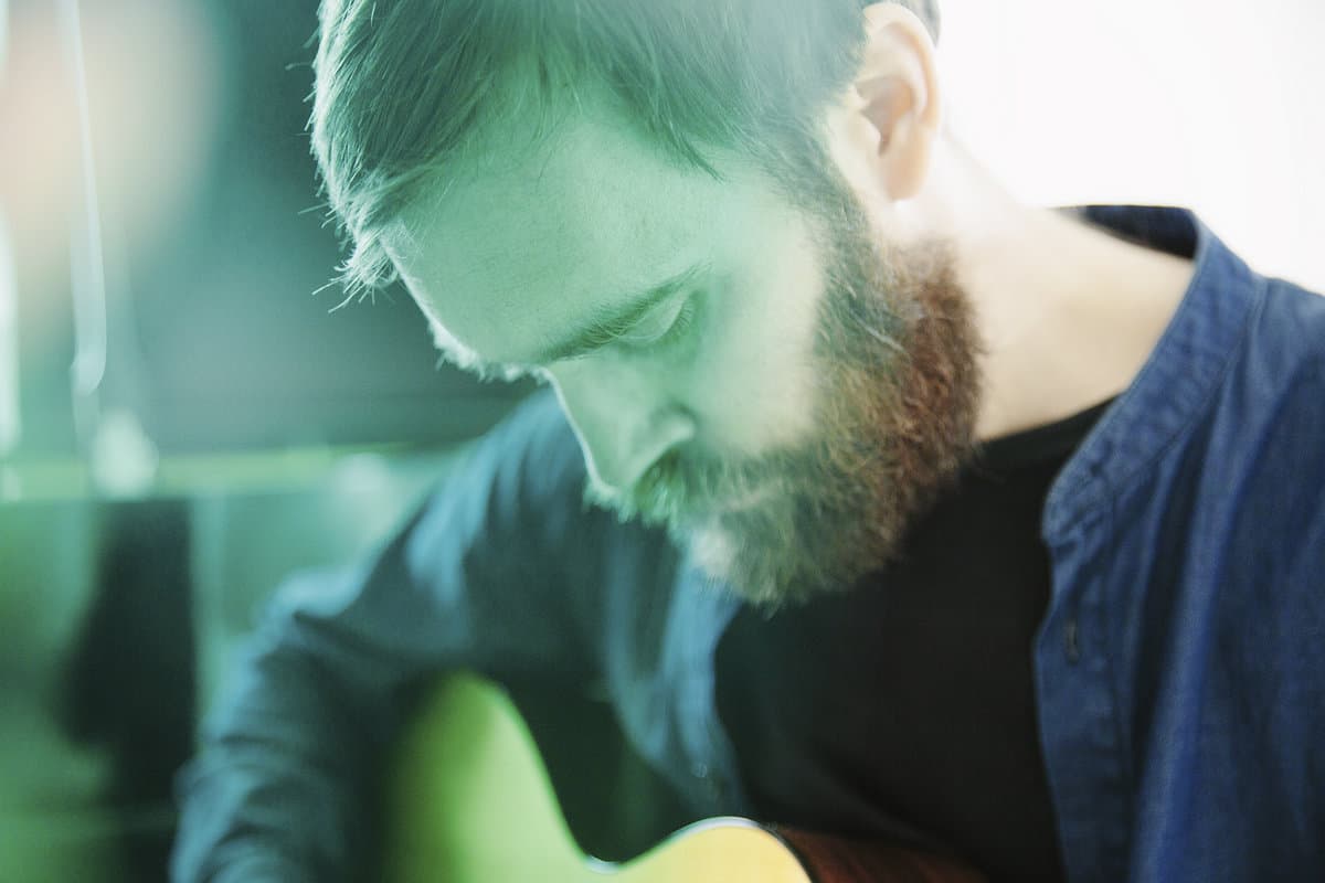 A man with a beard and closed eyes is playing a guitar, with soft lighting creating a tranquil atmosphere.