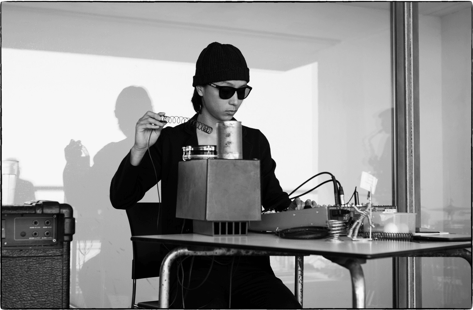 A person wearing sunglasses and a beanie is sitting at a table with audio equipment, holding a coil spring, and appears to be engaged in a sound setup.